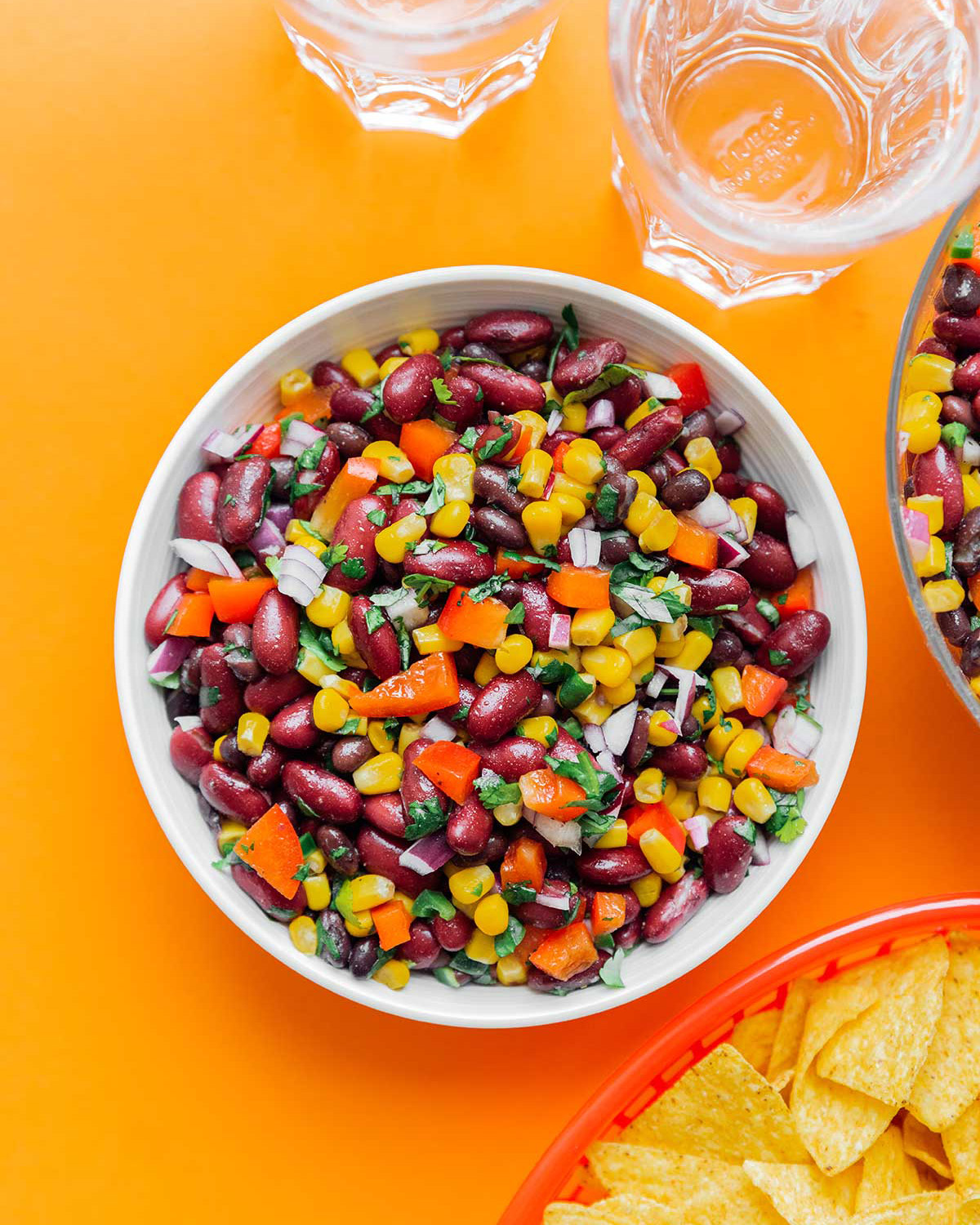 A bowl of bean salad sits on an orange table with chips to the side. This is great to make for summer RV recipes.