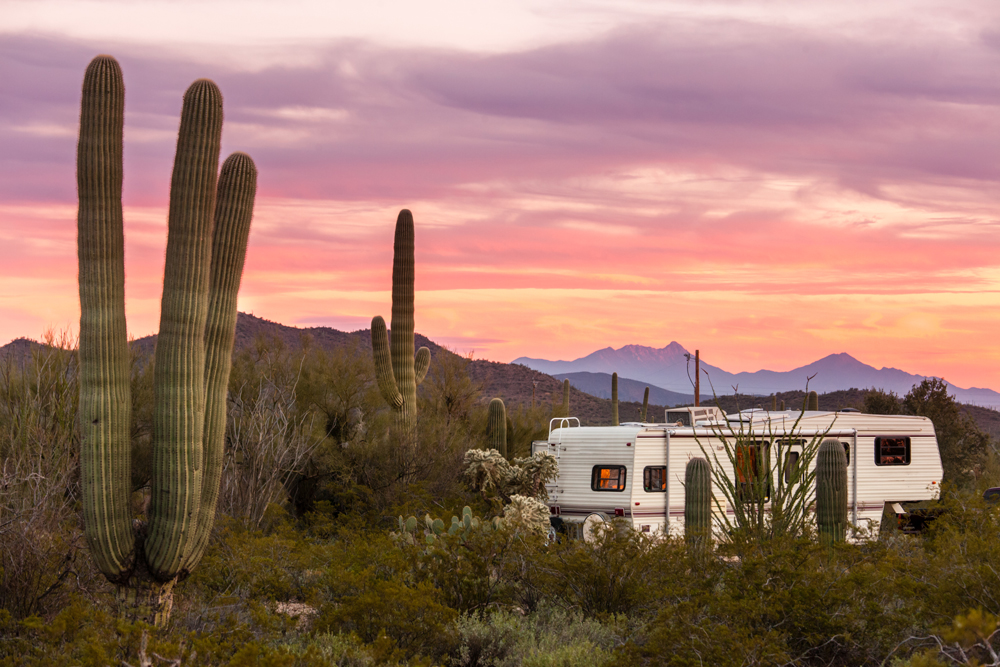 An older fifth wheel is parked among cacti at sunset in the desert.